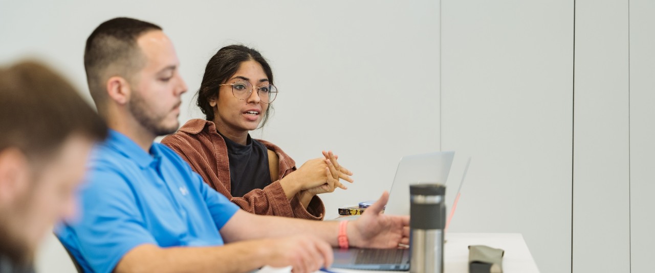 three students, one blurred in the foreground, one with a blue shirt in the middle ground, and one in a black and brown top clearest in the background, in a Lindner classroom