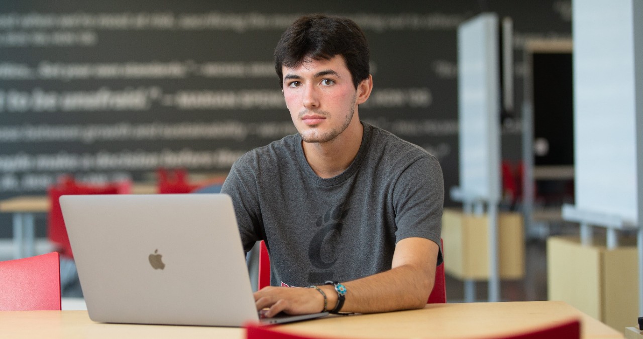 A male student sits at a table with a laptop.