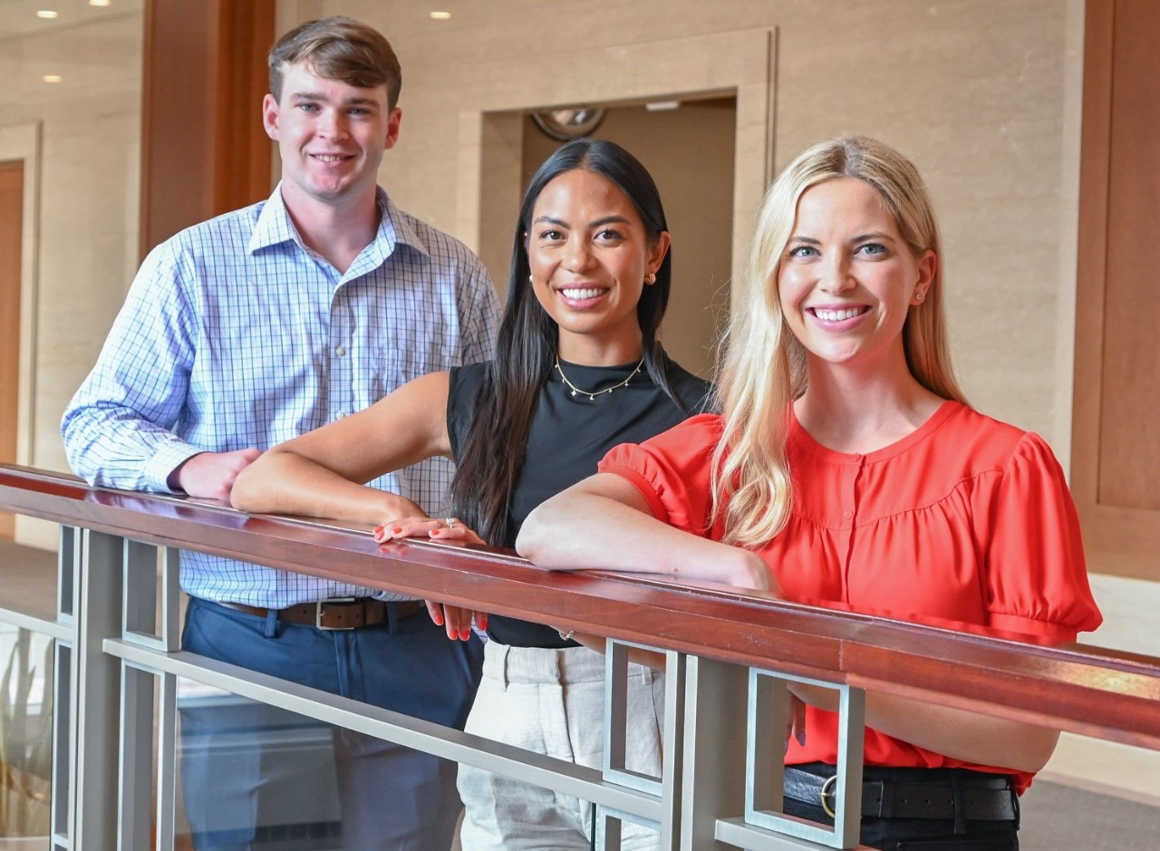 Two women and one man pose in professional dress by leaning on a railing.