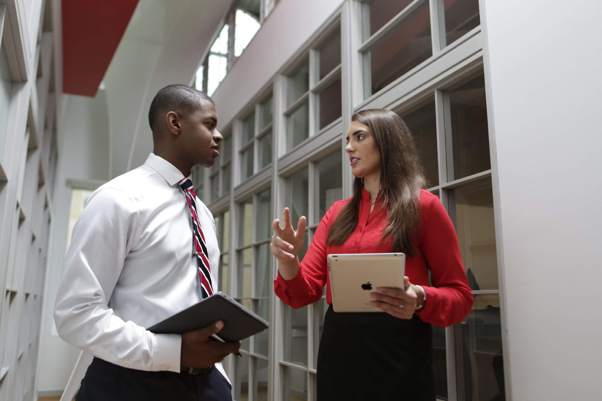 Professionally-dressed man and woman conversing in a hallway, holding tablet computers