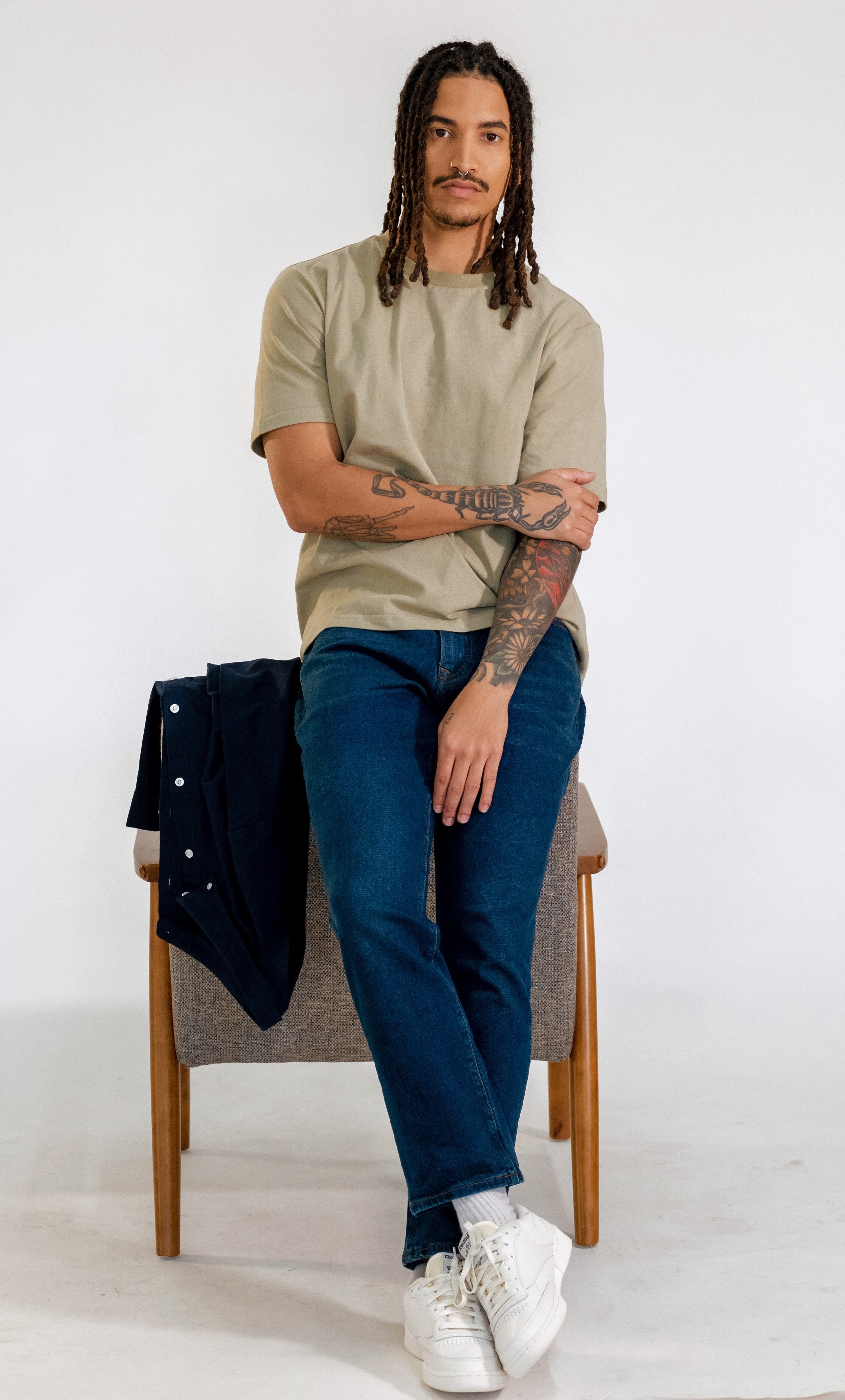 A clothing model sits on a wooden stool with a tan shirt and blue jeans on.