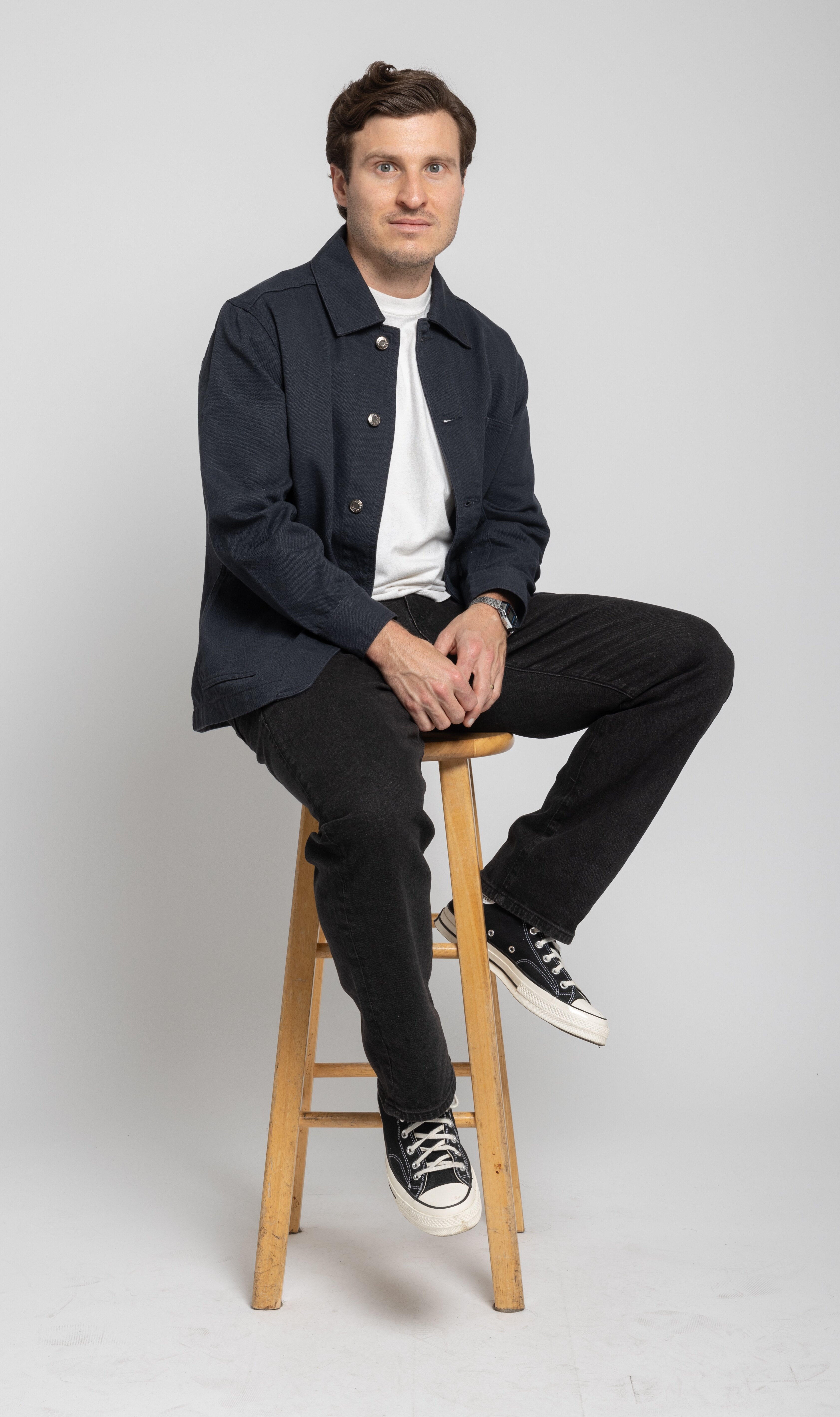 Mike Monnette sits on a wooden stool in a dark jacket and pants and a white shirt