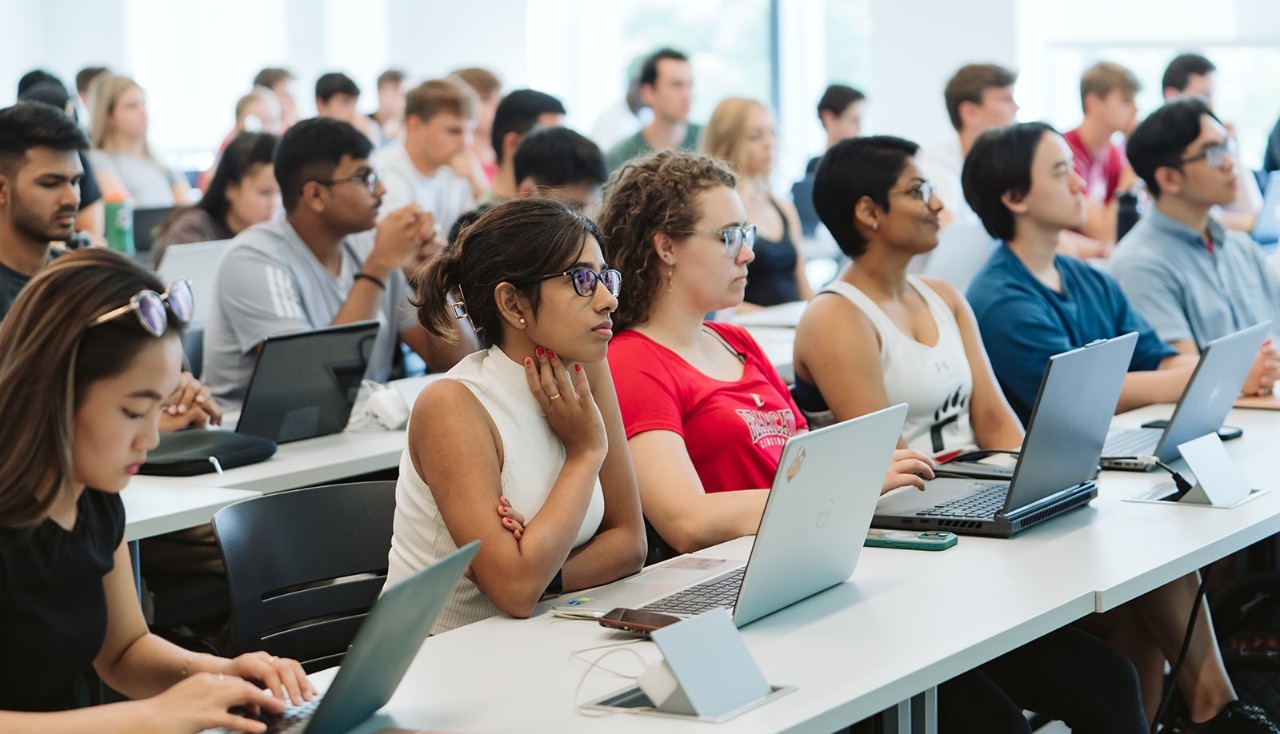 Students sit in rows, looking forward with their laptops open.