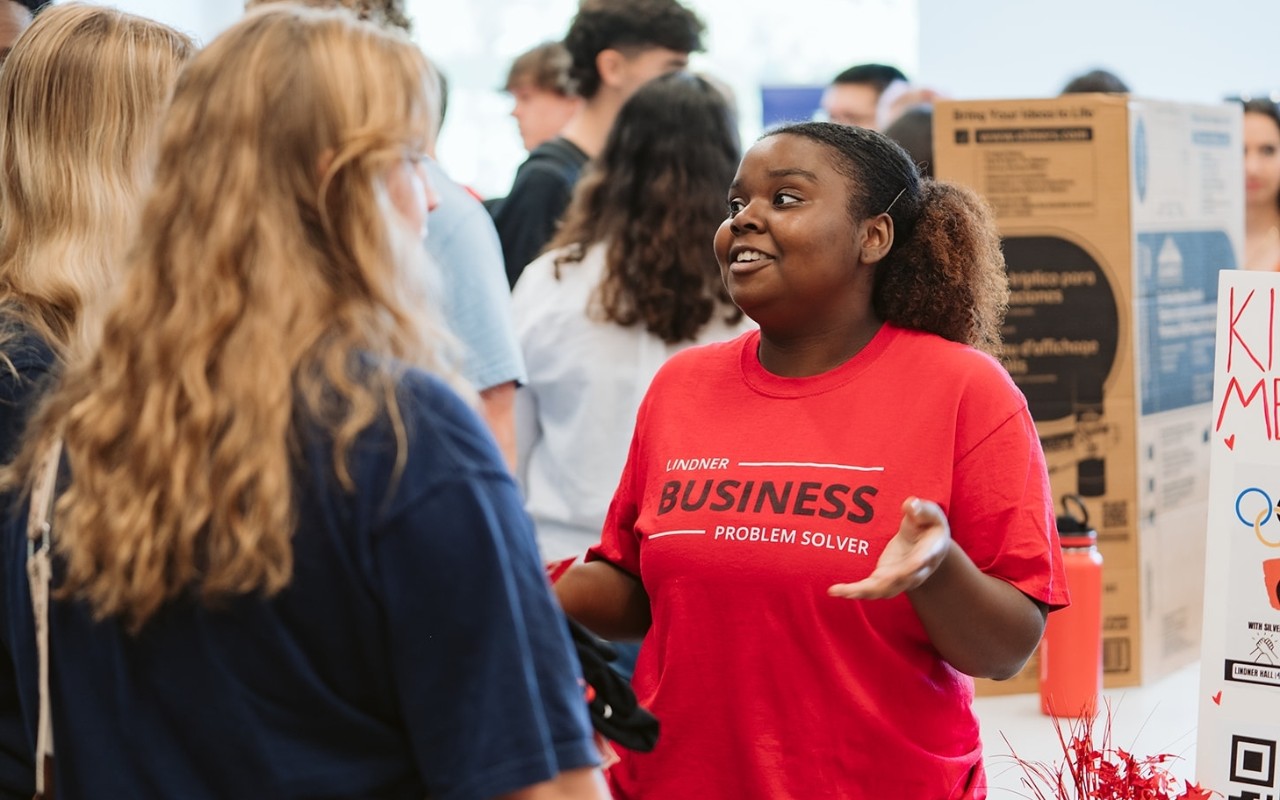 A Lindner student in a red shirt speaks to two other students at an involvement fair.